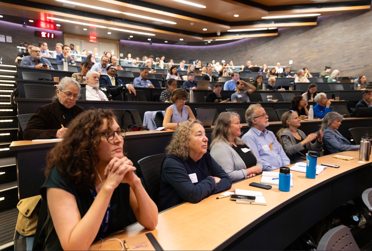A large group of men and women of varying demographics can be seen attending a symposium in an amphitheater-style auditorium.