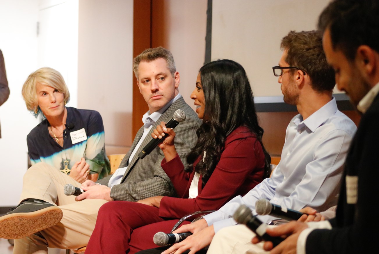 A tight crop of a female speaker in a red suit holding a microphone while surrounded by four other panelists.