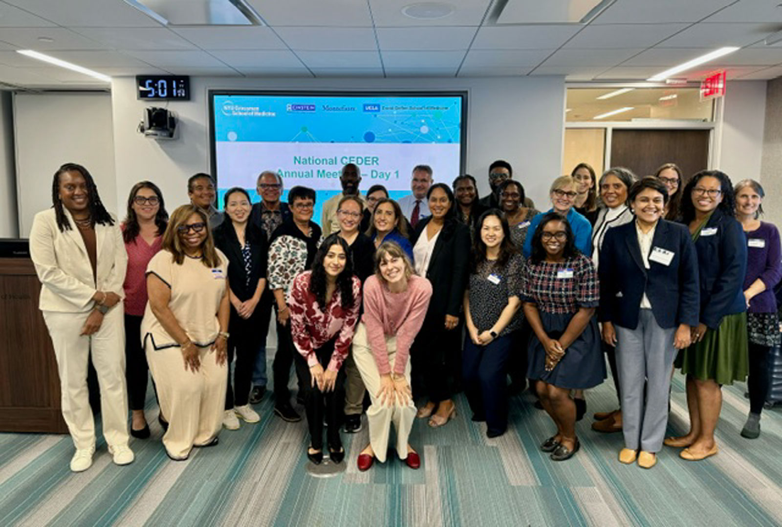 A group of people standing in front of a video board in a conference room.