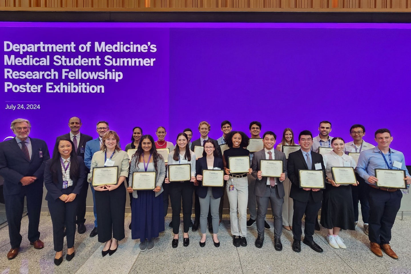 A group of people stands while holding certificates in front of a sign stating Department of Medicine’s Medical Student Summe Research Fellowship Poster Exhibition.