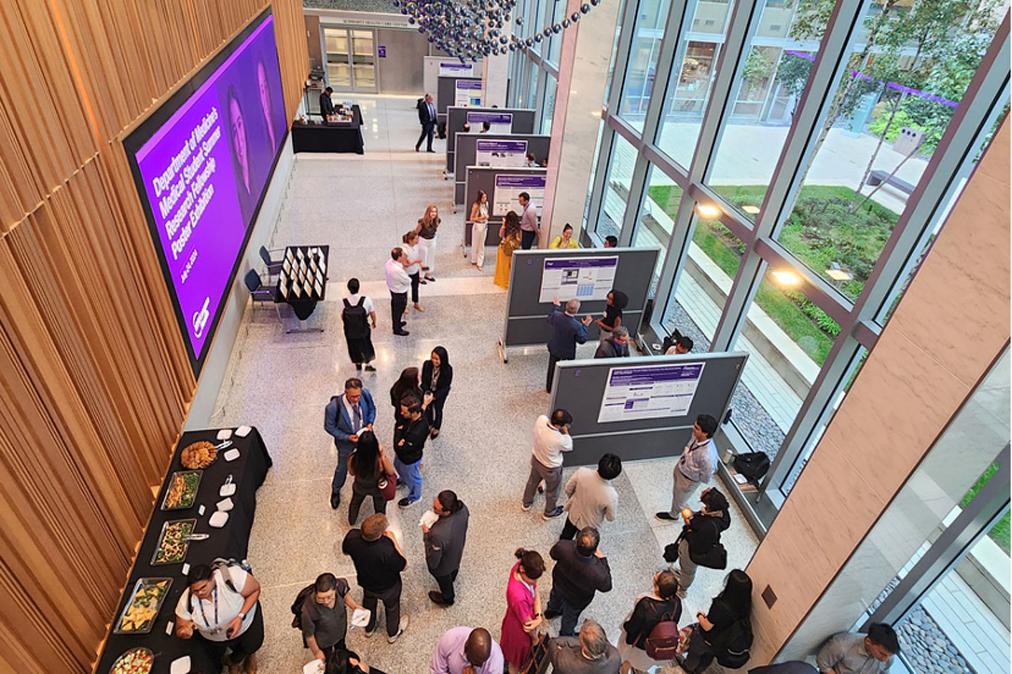 A large group of people gathers in an atrium with floor-to-ceiling windows and research posters mounted on display boards.