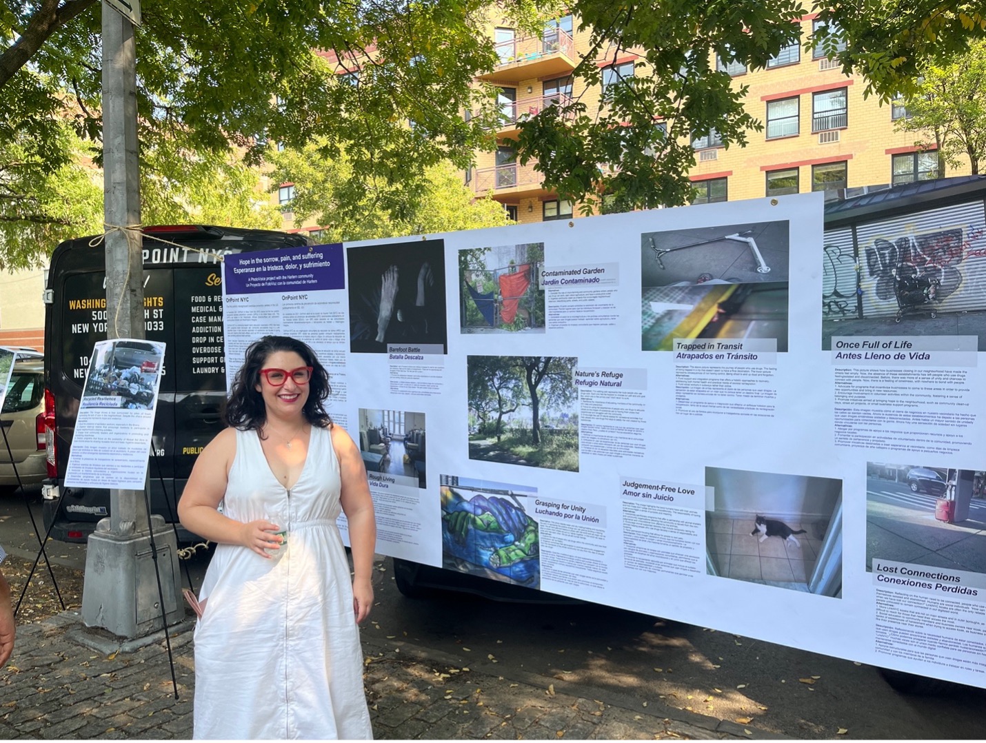 A woman in a white dress and red glasses stands next to a banner displaying photos and text descriptions of the images.