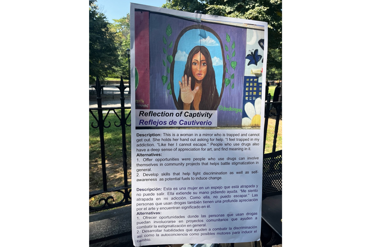 Photo displayed on a fence in the park shows a colorful painted mural of a woman with long hair and one palm facing out.