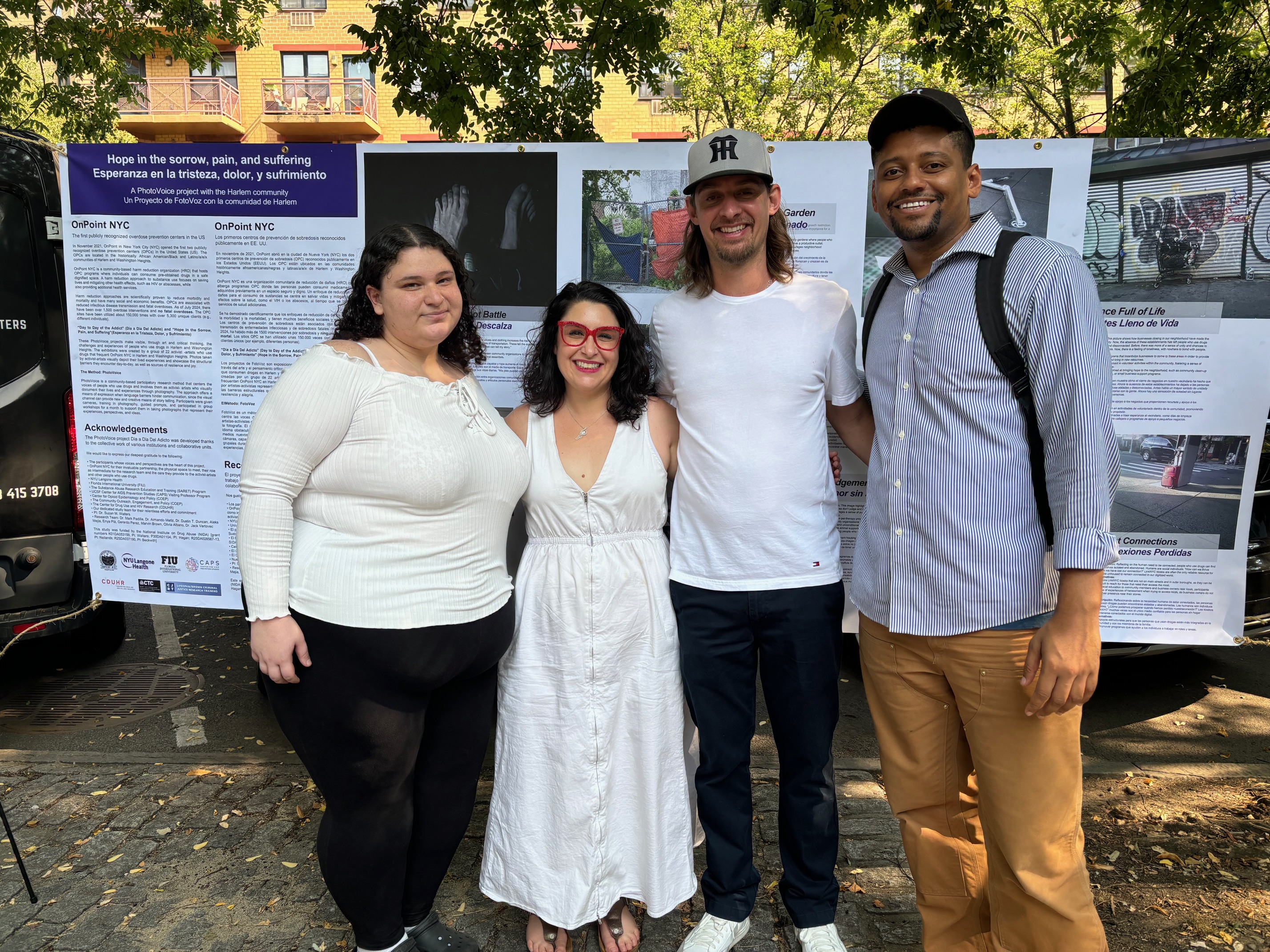 Group of people standing in front of informational posters outside under a tree.